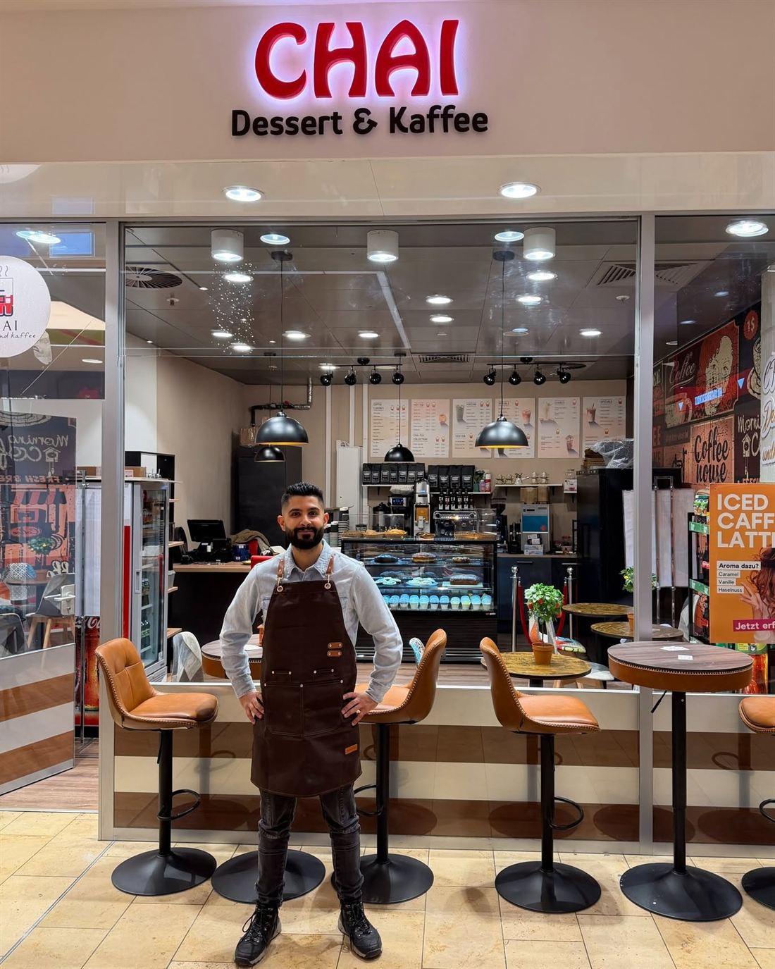 Teammitglied in brauner Schürze vor dem Laden CHAI Dessert & Kaffee mit Theke und Barhockern