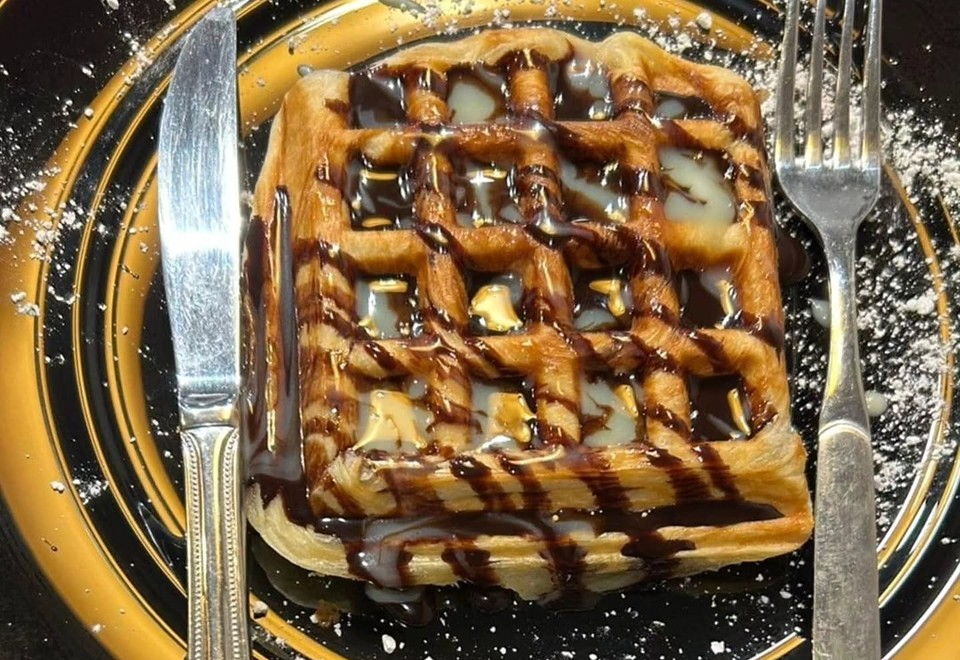 Croffle mit Schokoladen- und Vanillesauce und Puderzucker auf einem schwarzen Teller